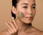 beautiful naked asian girl massaging face with jade roller isolated on beige